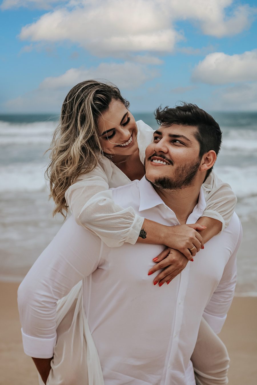 A smiling couple on the beach, she is on his back with her arms around him